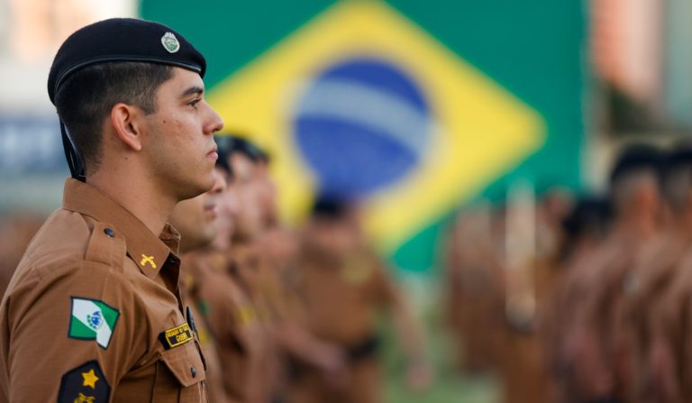 Policiais militares do paran&aacute; perfilados com a bandeira do brasil no fundo
