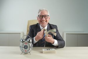 Um homem sentado à uma mesa, contando dinheiro e sorrindo. Sobre a mesma estão um relógio e várias notas empilhadas.